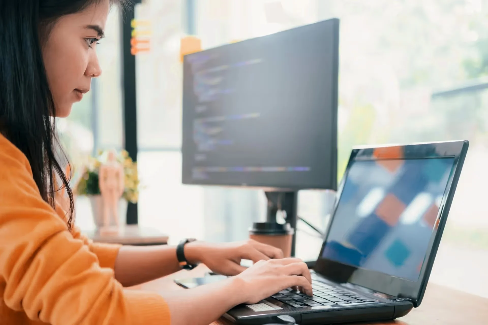 Person in an orange sweater typing on a laptop, with code visible on a large monitor nearby.
