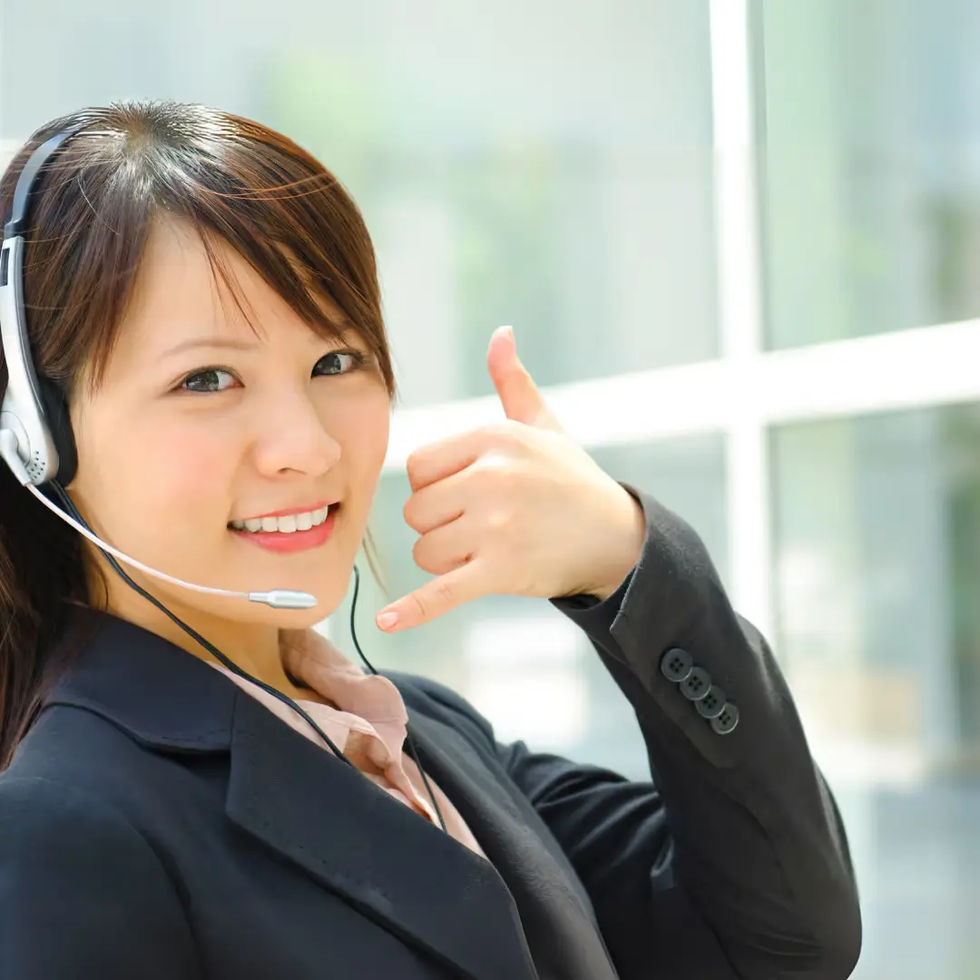 A woman wearing a headset, dressed in a blazer, smiles and gestures with her hand in a phone call symbol.