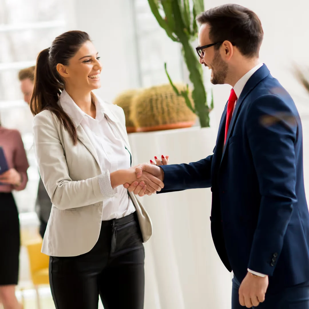 A man and a woman in business attire are shaking hands in an office setting, with a potted plant in the background.