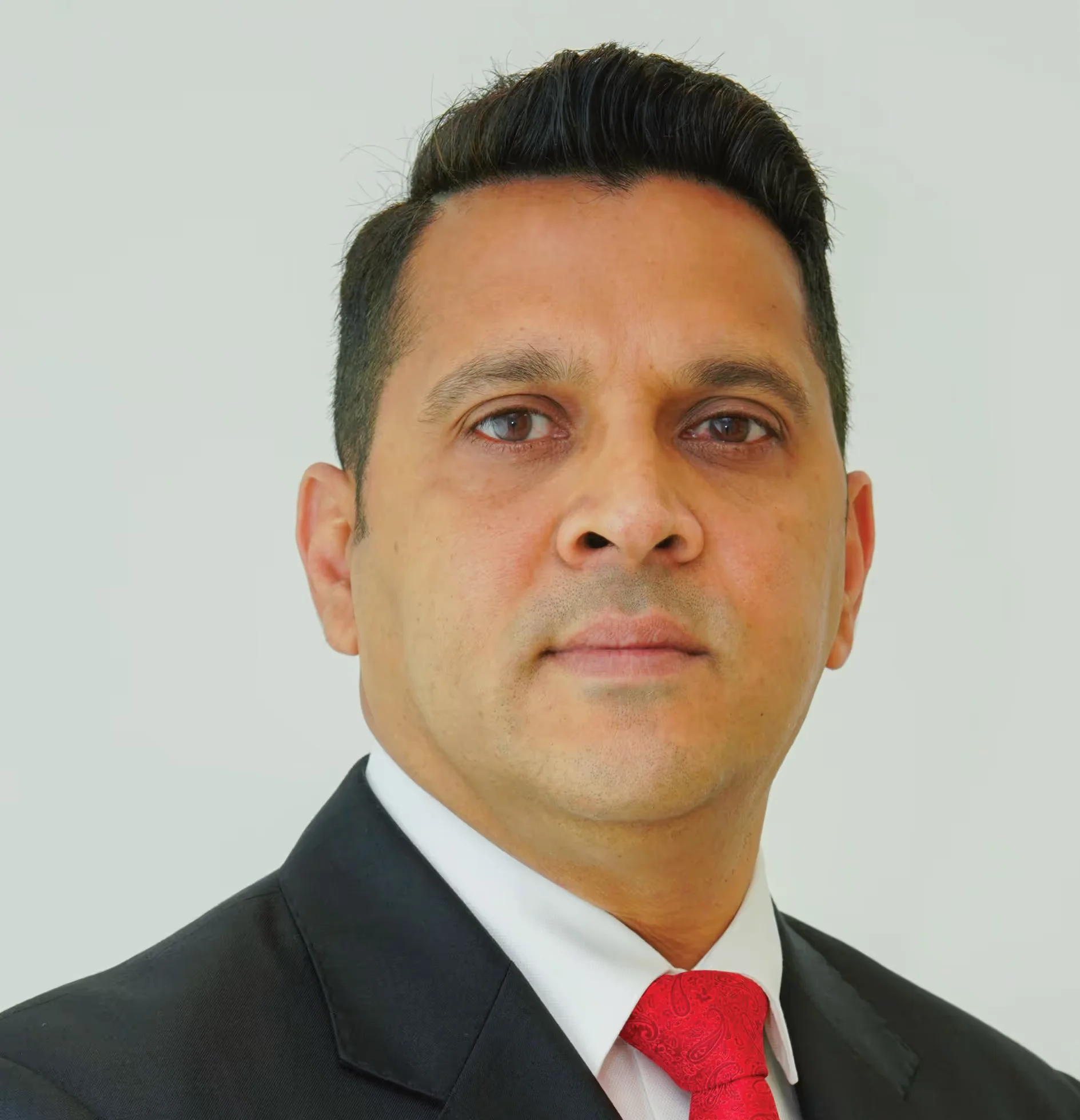A man in a black suit with a red tie, looking straight ahead against a plain background.