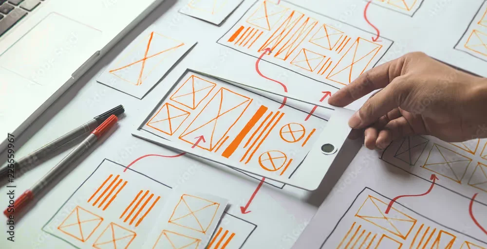 A person holds a tablet over wireframe sketches on paper, showing website layout designs with pens nearby.